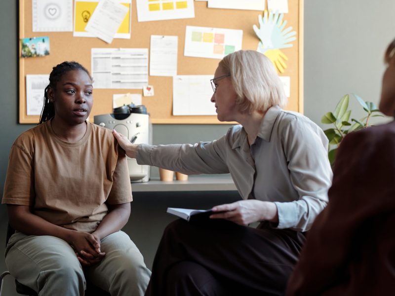 Blond mature female psychologist touching shoulder of upset African American girl while comforting her at psychological session