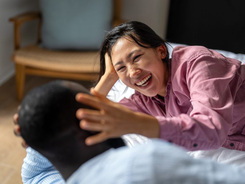 Excited child-free diverse, multiethnic couple embracing and laughing in modern living room, calm lifestyle